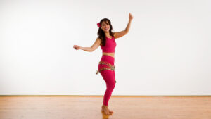 Shlomit Oren, wearing a bright pink belly dance outfit with gold accents dances barefoot on a wooden floor dancing for anxiety relief.