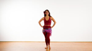 Shlomit Oren in maroon leggings and a matching top stands barefoot on a wooden floor with her hands on her hips, wearing a colorful scarf around her waist and a flower in her hair ready to belly dance for balancing the sacral chakra.