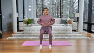 A smiling woman, Melissa Gutierrez, in mauve workout clothes sits on a chair atop a pink yoga mat in a modern living room, holding her hands on her stomach demonstrating how to activate the core for awareness.