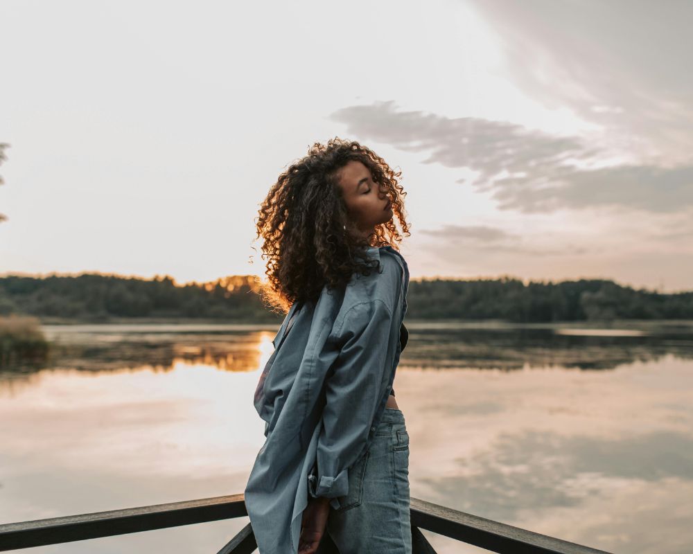 An African American woman with curly hair stands on a wooden deck by a calm lake at sunset, with eyes closed and head tilted back, surrounded by trees and a cloudy sky.