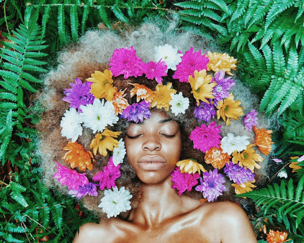 An African American woman lying on grass with flowers in her hair practicing mindful self-love outdoors