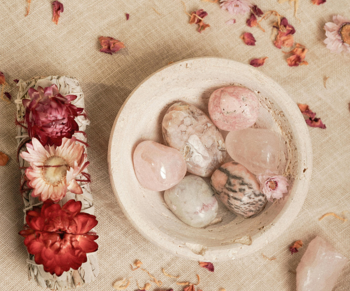 Healing crystals including Rose Quartz set in a bowl on a table with scattered flower petals. There is also a small plate with three red and pink flowers on the side.