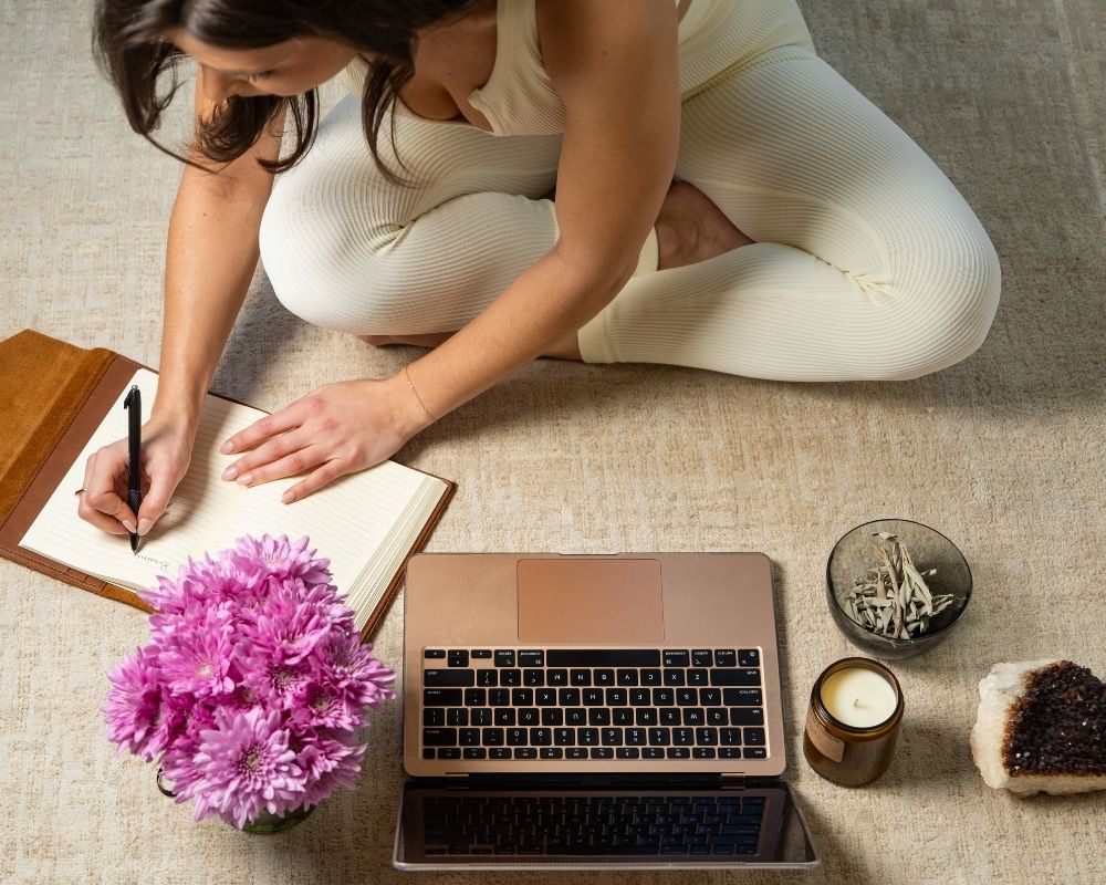 A woman working on a computer beside sage and a journal, representing the balance between artificial intelligence and inner wisdom.