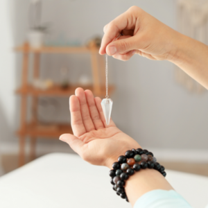 Maria Marshall demonstrating how to work with a pendulum. She is lifting a clear quartz pendulum with one hand and holds it over her open palm. Dressed in white and seated, she is serene surrounded by plants.