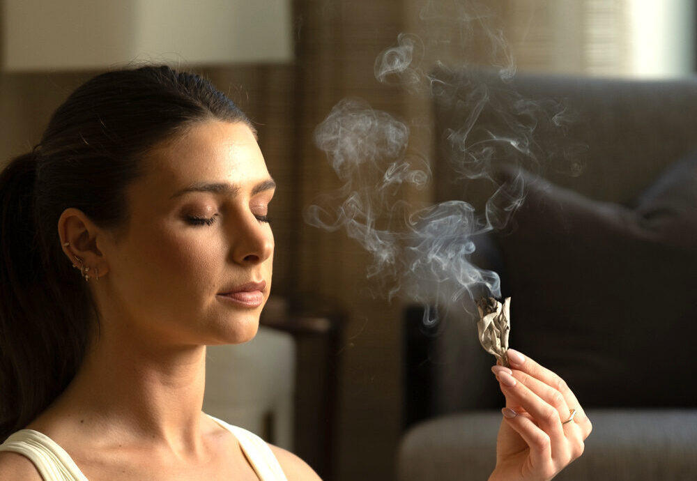Woman at home, sitting in yoga pose doing the energy clearing clearing practice of saging. She is holding a lit sage bundle in her hand and letting the smoke from the sage bundle surround her face as she sages herself.