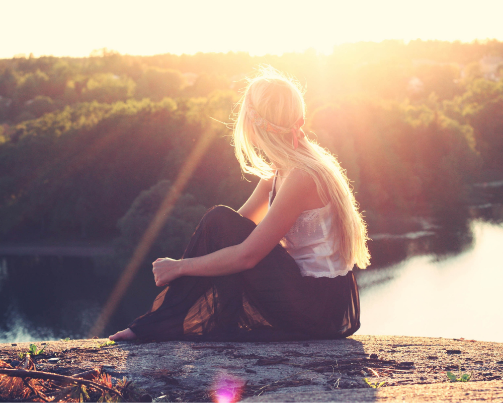 A woman with long blonde hair sits on a rock overlooking a river and forest at sunset, with sunlight creating a warm, glowing effect. She takes in the beauty of Gaia on earth day.