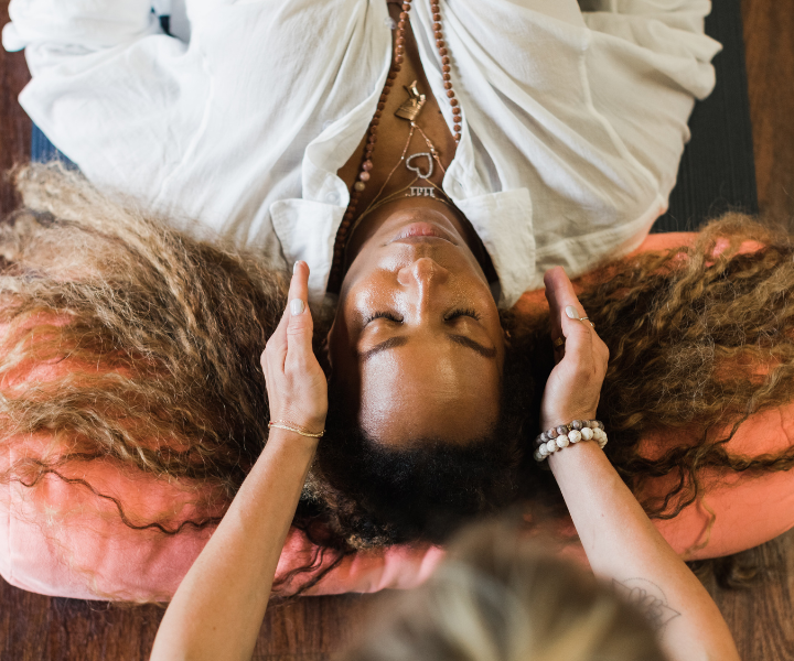 An African American woman with long curly hair lies on a pink cushion, eyes closed, as another gently places hands on their head performing reiki—an image capturing Maria Marshall guiding a soothing practice for the Master Level Reiki Certification.