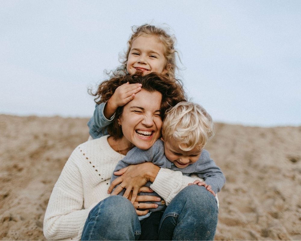 A woman sits on the sand at the beach, smiling and hugging two young children. One child hugs her from behind, smiling, while the other child sits on her lap, looking down the representation of motherhood.