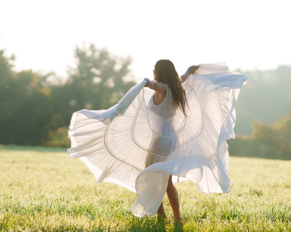 A Gaia Goddess woman in a flowing white dress stands in a sunlit field, holding the dress out wide as it billows in the breeze where she celebrates the Summer Solstice.