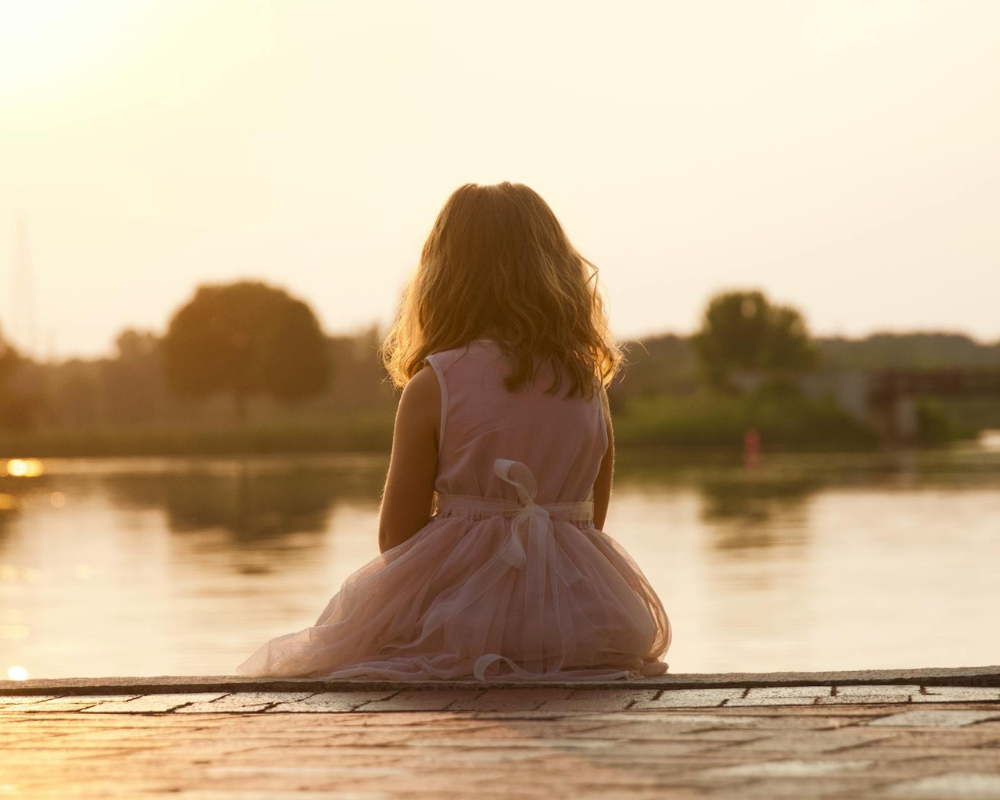 A young girl in a pink dress sits on the edge of a dock, facing a calm lake at sunset. Trees and distant shoreline are softly blurred in the warm, golden light as she sits sadly pondering emotions.