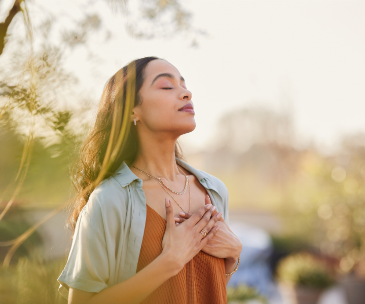 A woman stands outdoors with her eyes closed, hands gently resting on her chest, embodying the calm and peaceful essence of the August 2026 Monthly Healing and Breathwork. She wears a light shirt over a brown top, surrounded by sunlight and greenery that enhance the serene atmosphere.