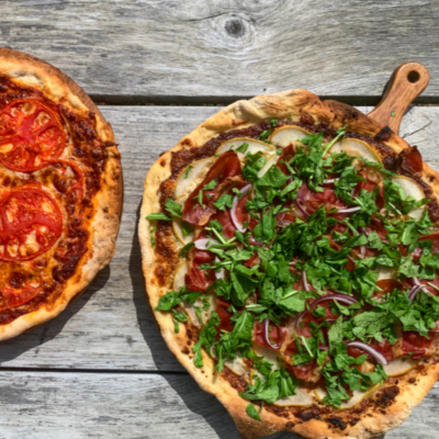 Two homemade pizzas on a wooden table capture the inviting spirit of the Gaia Goddess Women’s Reiki Certification Levels 1&2 Retreat; one is topped with sun-ripened tomato slices and melted cheese, while the other bursts with vibrant greens, red onion, and crispy bacon.
