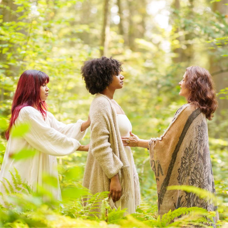 Three women stand in a lush, green forest. Two women gently hold the hands and shoulders of the third woman, creating a supportive, peaceful atmosphere surrounded by sunlight and foliage—a scene reminiscent of the connection nurtured at a Reiki 1 & 2 Certification Retreat.