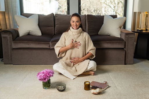 Maria Marshall, Gaia Goddess founder and CEO, sits cross-legged on a rug in front of a couch, smiling with one hand on her chest and one on her belly. Nearby are pink flowers, a candle, crystals, a journal, and a bowl, suggesting a peaceful, meditative setting.