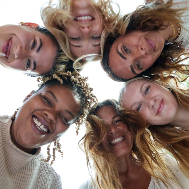 Six women form a circle, their heads together and faces beaming with joy as they look down at the camera, radiating unity and friendship that beautifully captures the essence of the Gaia Goddess Women’s Reiki Certification Levels 1&2 retreat. The background is bright and white.