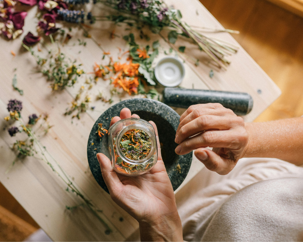 A woman holds a small glass jar filled with dried herbs over a table scattered with colorful dried flowers and stems, preparing or arranging the herbs with their other hand.
