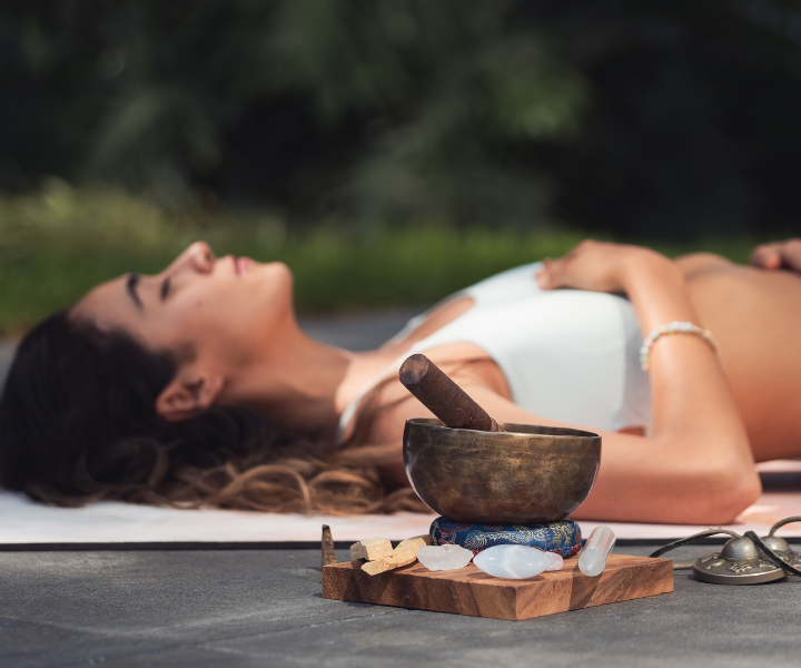 A woman laying down in nature hands on chest behind tuning bowl and crystals laid out in a circle as she engages in breathwork.