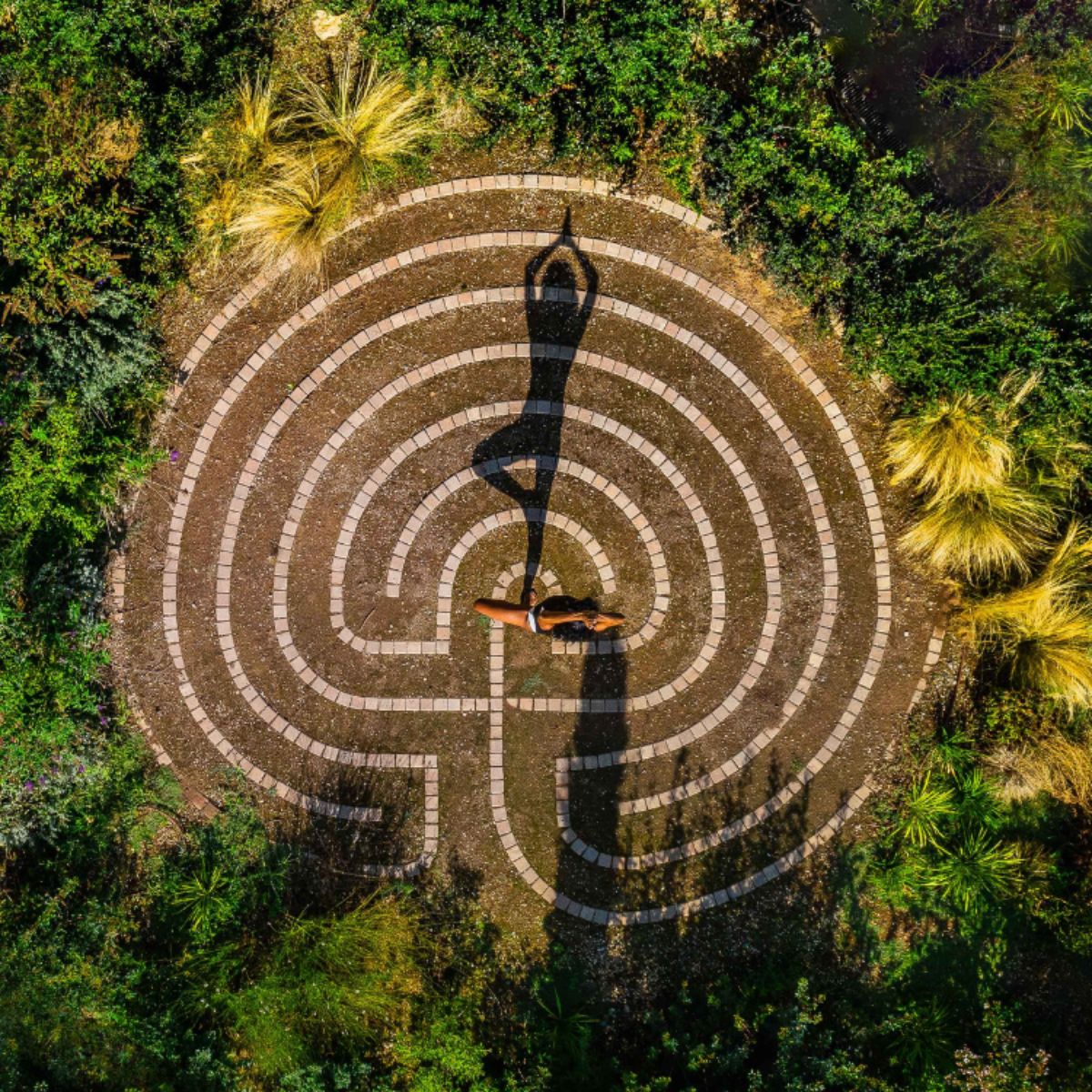 A person stands in a yoga pose in the center of a circular stone labyrinth surrounded by greenery.