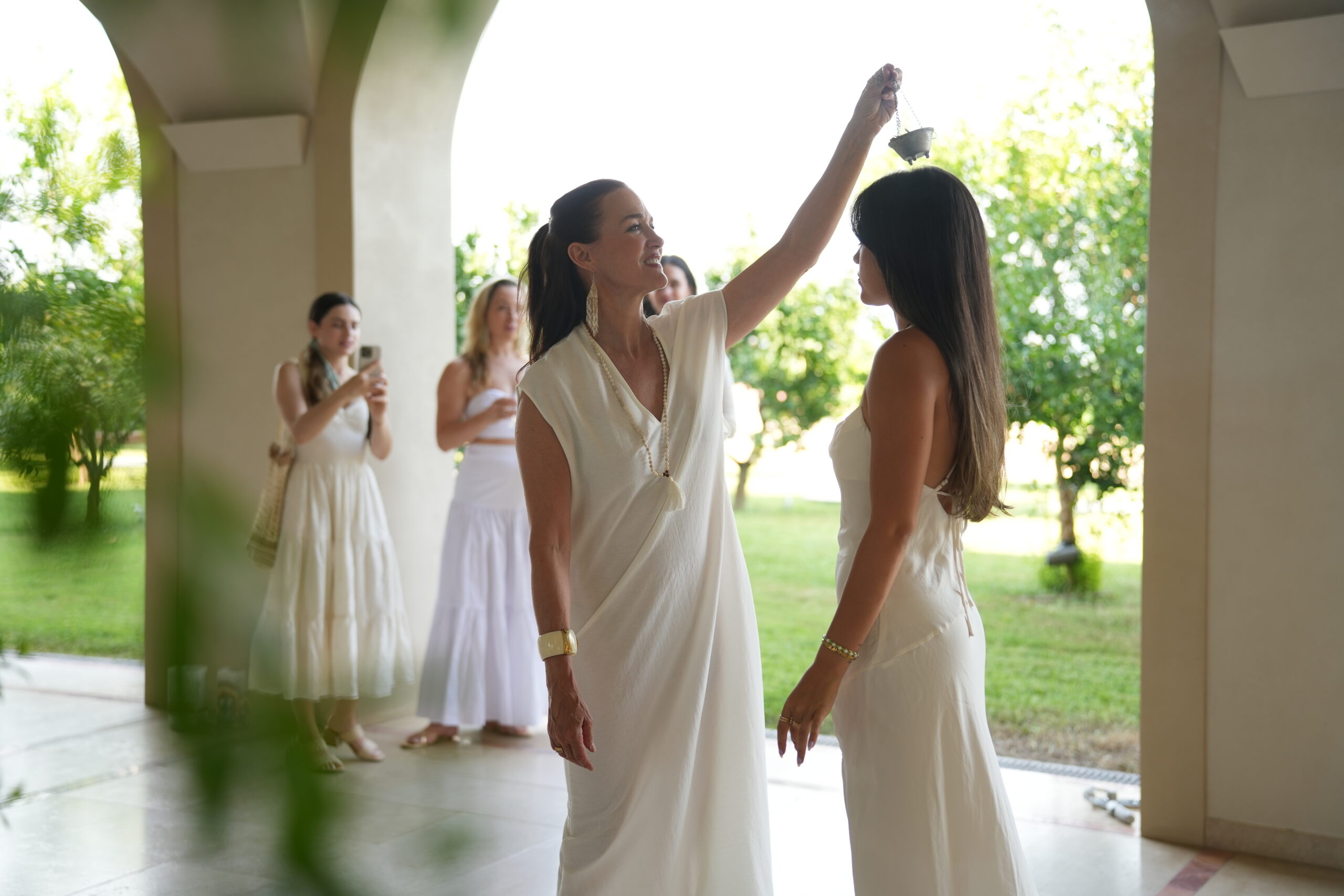 A woman in a white dress holds a small incense burner over the head of another woman in a white dress, while two other women in white dresses watch in the background.