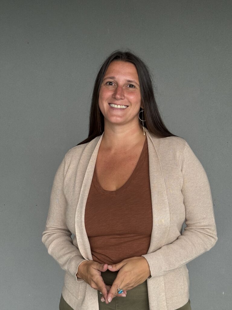 A woman with long dark hair smiles at the camera, wearing a brown top and a beige open cardigan against a gray wall.