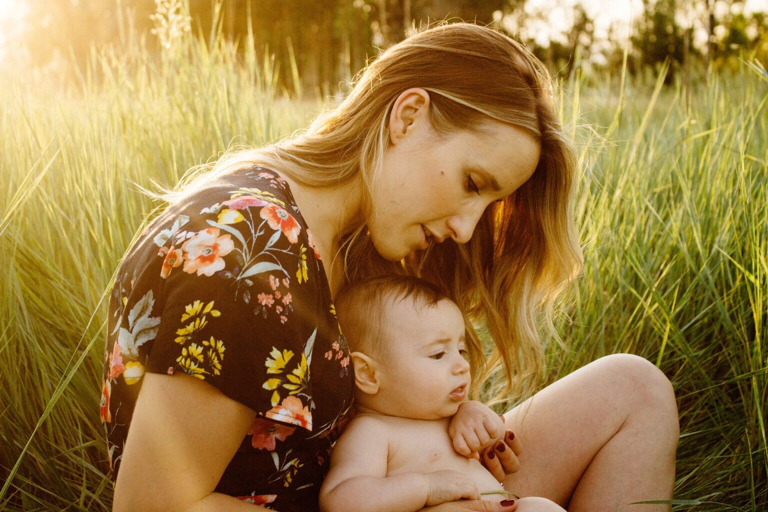 A mother sitting in a tall grassy field, holding her baby close.