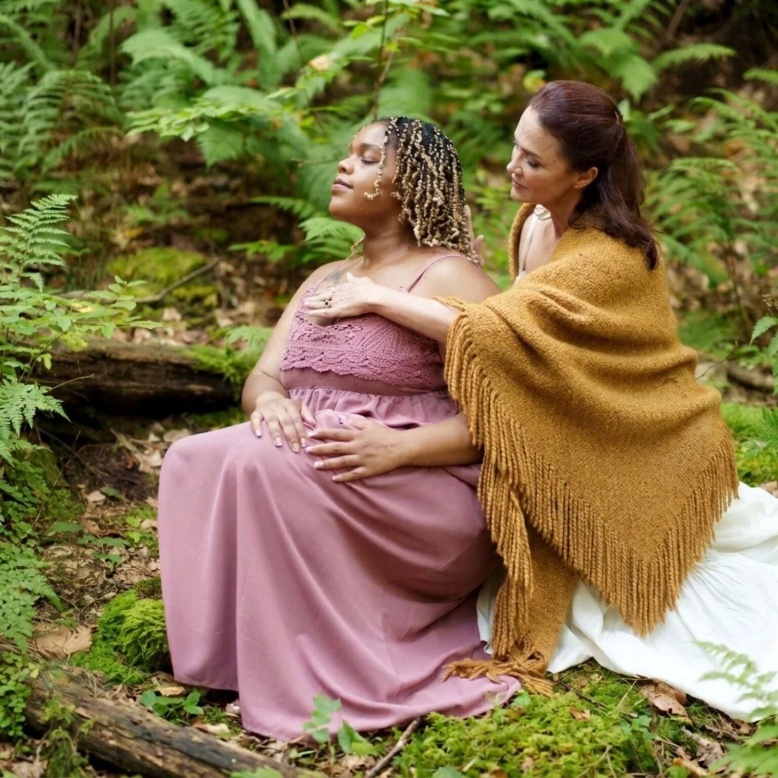 A woman in a yellow shawl places her hand on the chest of a pregnant woman in a pink dress as they sit together in a forest.