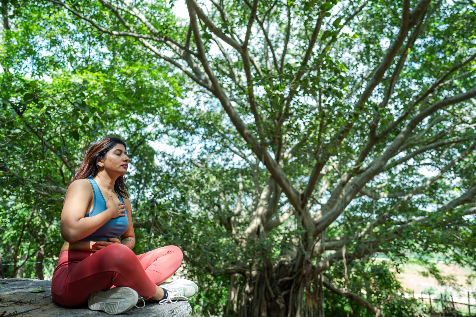 A woman in athletic wear sits in a meditative pose on a stone ledge under a large tree with her hand on her chest.