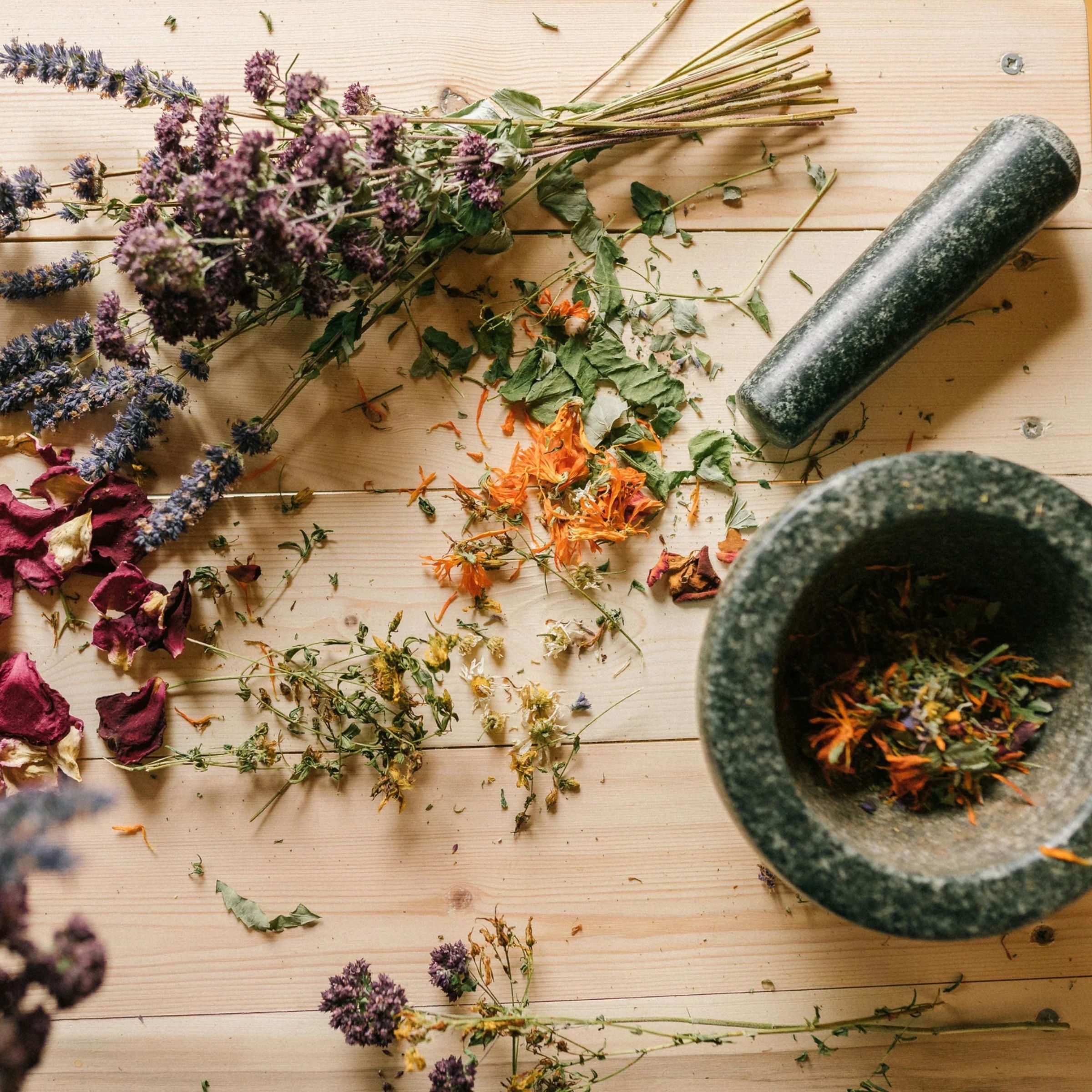 Dried herbs and flower petals scattered on a wooden table next to a stone mortar and pestle containing a herbal mixture.